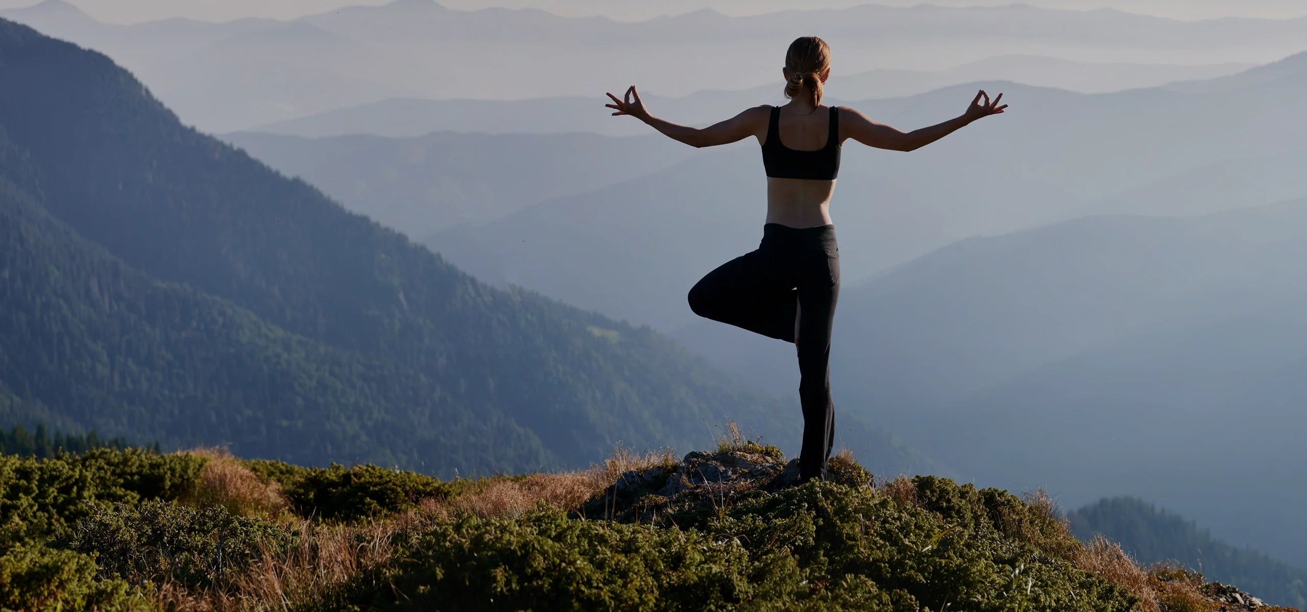 Eine Frau steht in Yoga-Pose auf einem Berggipfel und blickt in ein weites, nebliges Bergpanorama. Sie trägt schwarze Sportkleidung und hält beide Arme entspannt ausgestreckt. Die Szene strahlt Ruhe, Ausgeglichenheit und Naturverbundenheit aus.